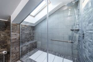 A contemporary bathroom with a skylight, featuring a spacious shower area enclosed by coloured glass screens. The shower area has a modern design with grey mosaic tiles and a sleek rain shower head, showcasing the use of coloured glass screens to enhance the bathroom's brightness.