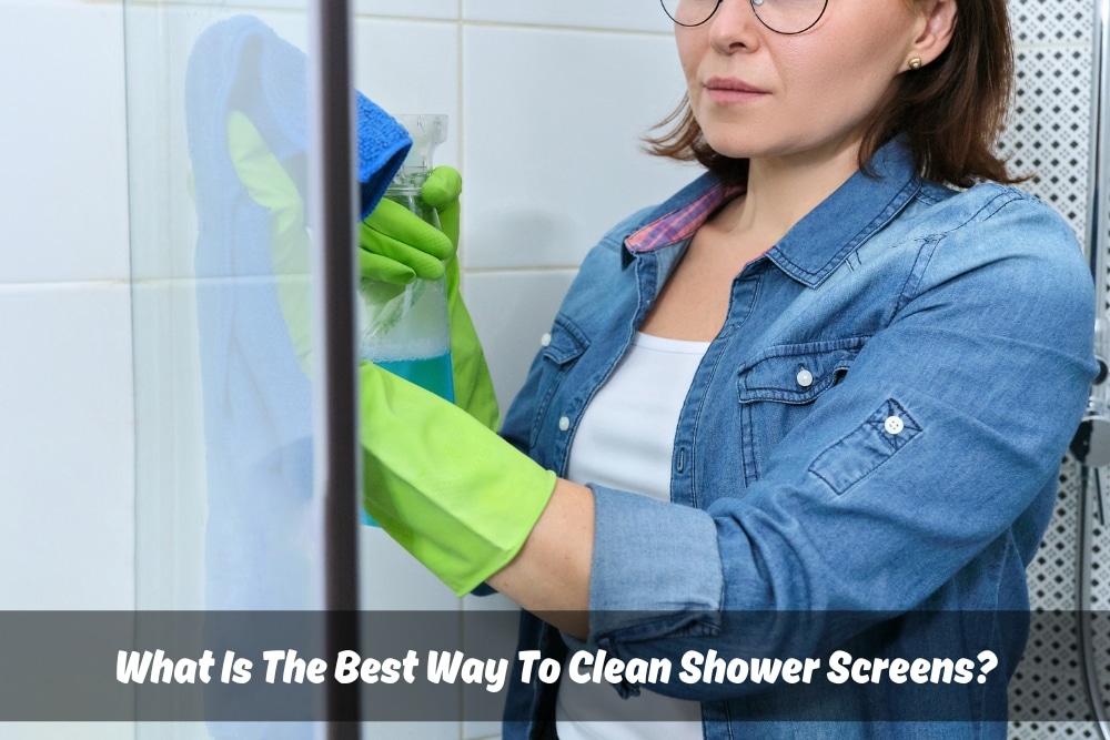 A woman wearing glasses and green rubber gloves is cleaning a shower screen with a blue cloth and a spray bottle of cleaning solution. This image illustrates the best way to clean shower screens effectively, highlighting the importance of using appropriate cleaning tools and protective gear.